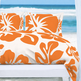 A bed dressed in a white duvet cover and matching pillowcases with a bold orange tropical floral pattern, set against a beach background with ocean waves and a white cabana.