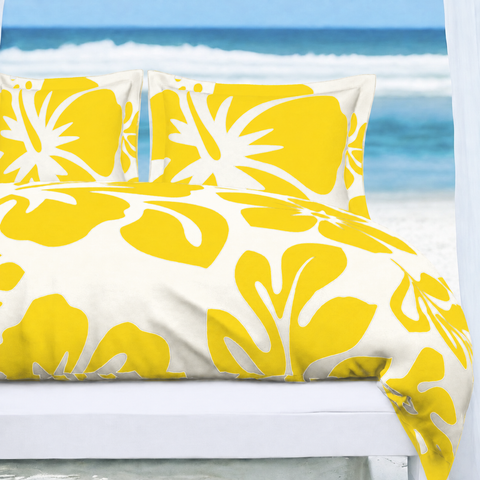 A bed dressed in a yellow and white tropical floral print duvet cover with matching pillowcases, set against a blurred beach background.